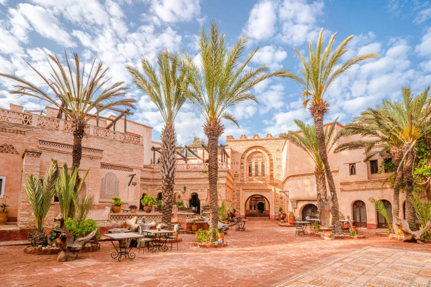 Moroccan courtyard with tall palm trees, terracotta walls and café tables under a blue sky