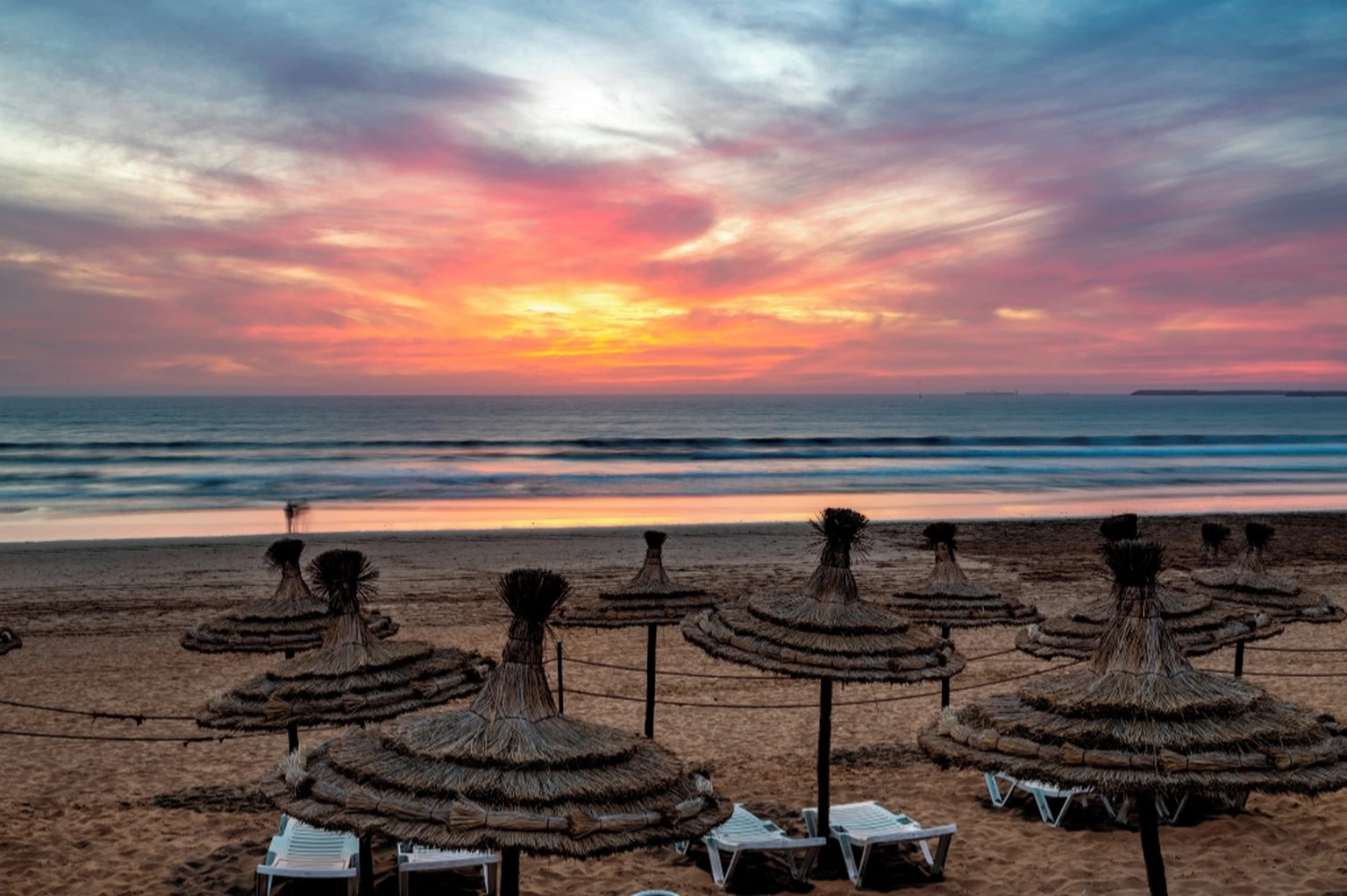 Dramatic sunset over Agadir beach with thatched umbrellas, ocean waves and colorful evening clouds
