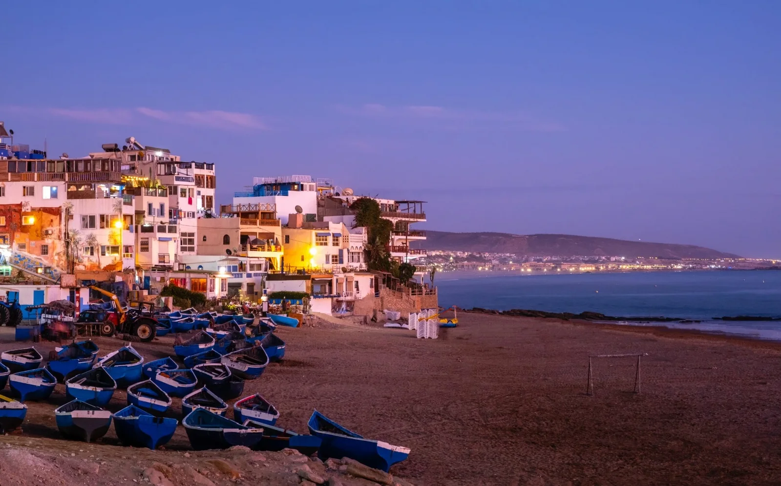 Taghazout bay at dusk — blue fishing boats on the sand below the lit village
